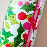 A close-up of a roll of wrapping paper featuring a festive holly and berry pattern in green, pink, and red on a white background.